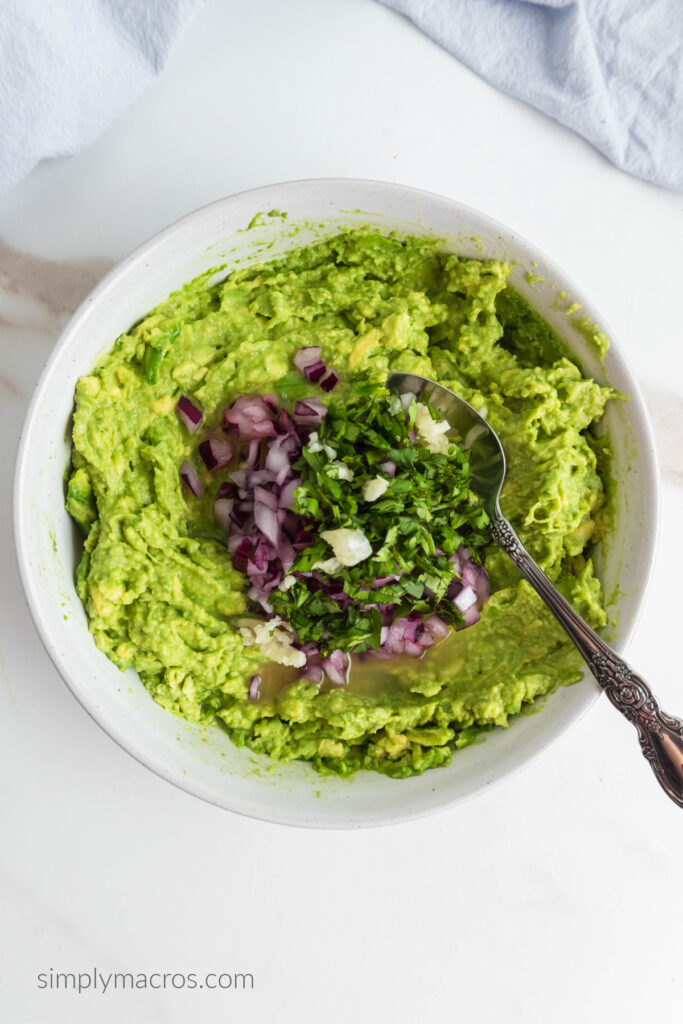 Mashed avocado, diced onion, minced garlic, and chopped cilantro in a white bowl, ready to mix into a homemade guacamole dip. 