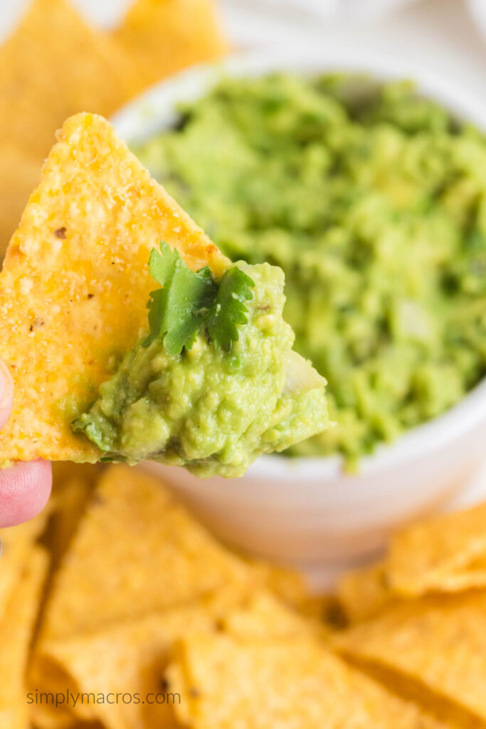 Close up shot of guacamole on a chip, with the bowl of guacamole and chips in the background. 