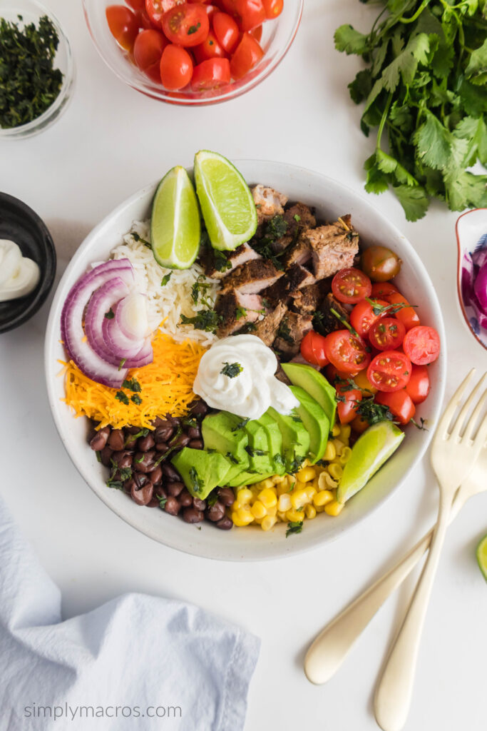 Overhead shot of a steak burrito bowl surrounded by chopped tomatoes, forks, cilantro, sour cream, and ready to eat. 