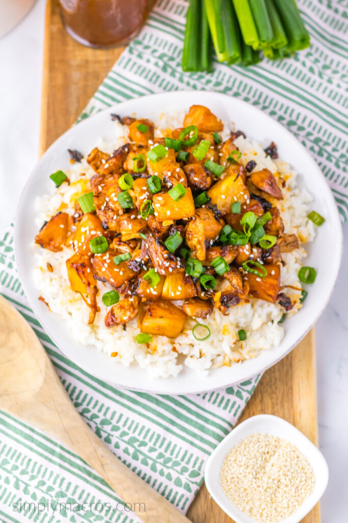 Brown sugar pineapple chicken served over a bed of rice on a white plate and topped with sesame seeds and chopped scallions. 