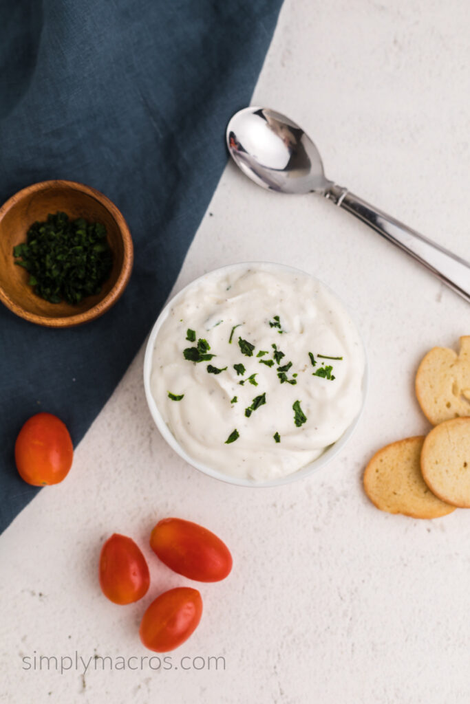 Cottage cheese ranch dip in a small bowl with fresh parsley sprinkled on top. Surrounded by tomatoes and bagel chips.