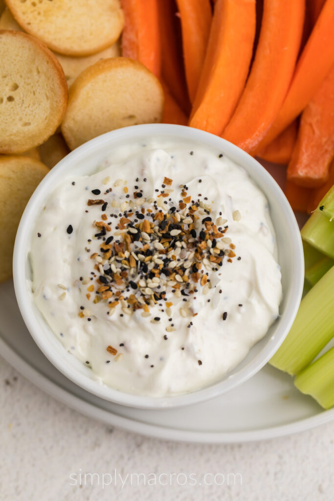 Everything bagel dip in a small bowl surrounded by bagel chips and vegetables. 