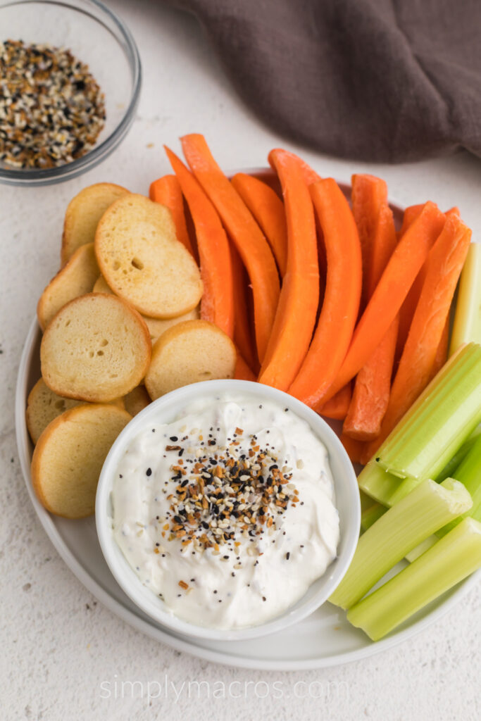 Everything bagel dip in a small bowl with seasoning garnished on top, surrounded by vegetables. 