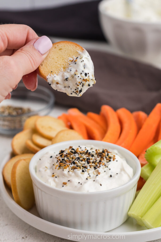 Everything bagel dip in a bowl surroundd by bagel chips and vegetables, and a bagel chip dipped and being held. 