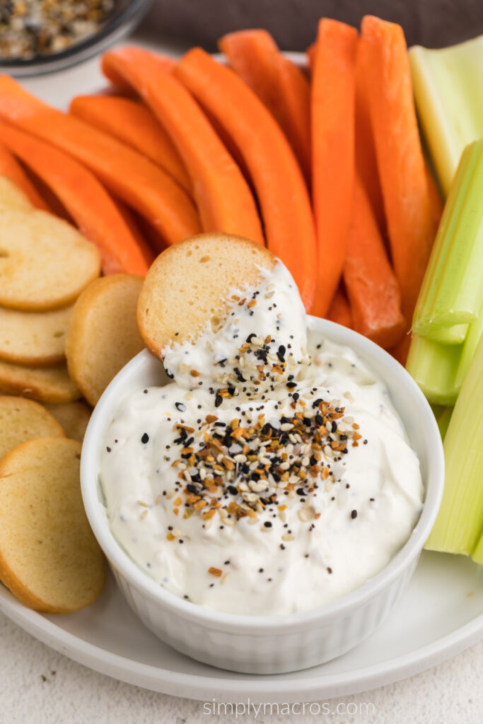 Everything bagel dip with a bagel chip in the dip, and surrounded by bagel chips and vegetables. 