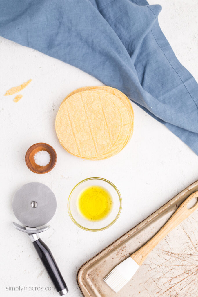 Corn tortillas, salt, and olive oil on a table with a basting brush, tray, and pizza cutter. 