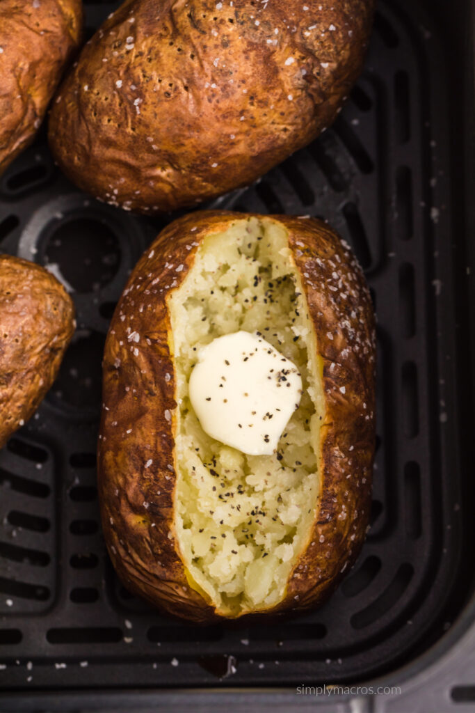 Baked potato in air fryer basket sliced open and topped with butter and pepper, ready to serve.