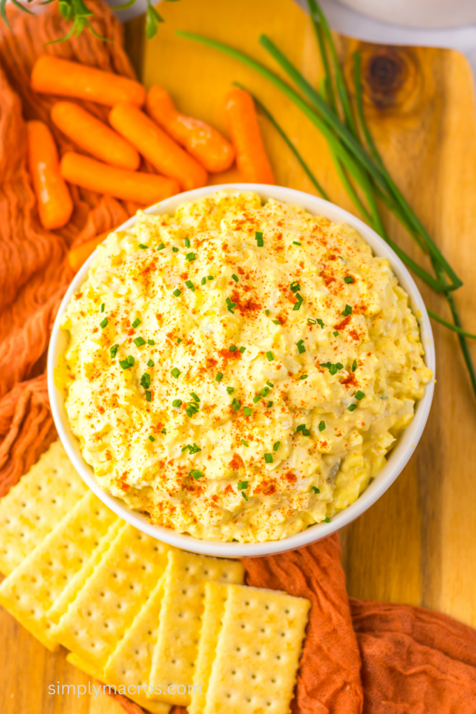 Deviled egg dip in a bowl, topped with paprika and chived and ready to serve with crackers and carrots on the side. 