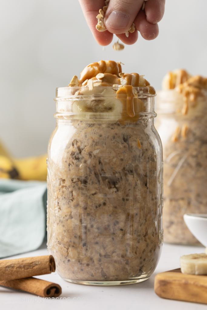 Peanut butter and banana flavored overnight oats in a jar with a hand sprinkling oats on top before serving. 