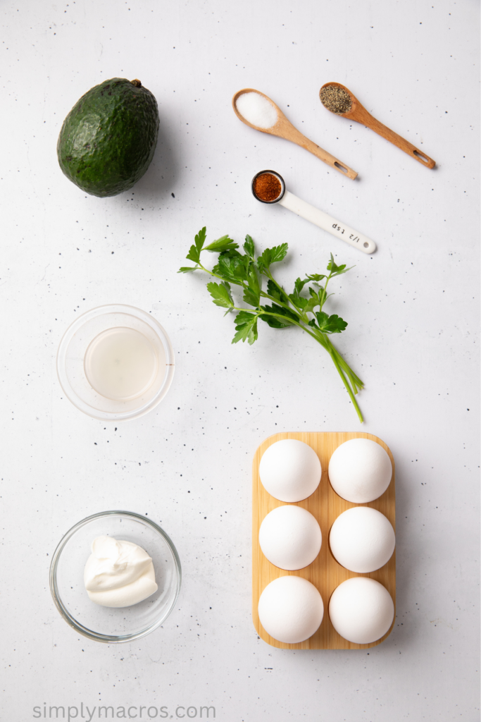 Ingredients on a table top, eggs, avocado, seasonings, lemon juice, mayo, and parsley. 